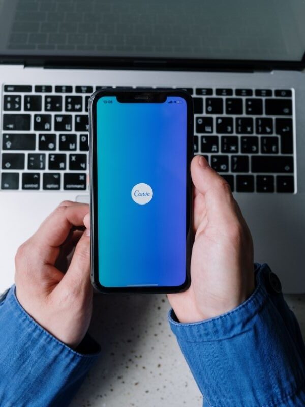 hand holding a phone with a Canva logo over a laptop.